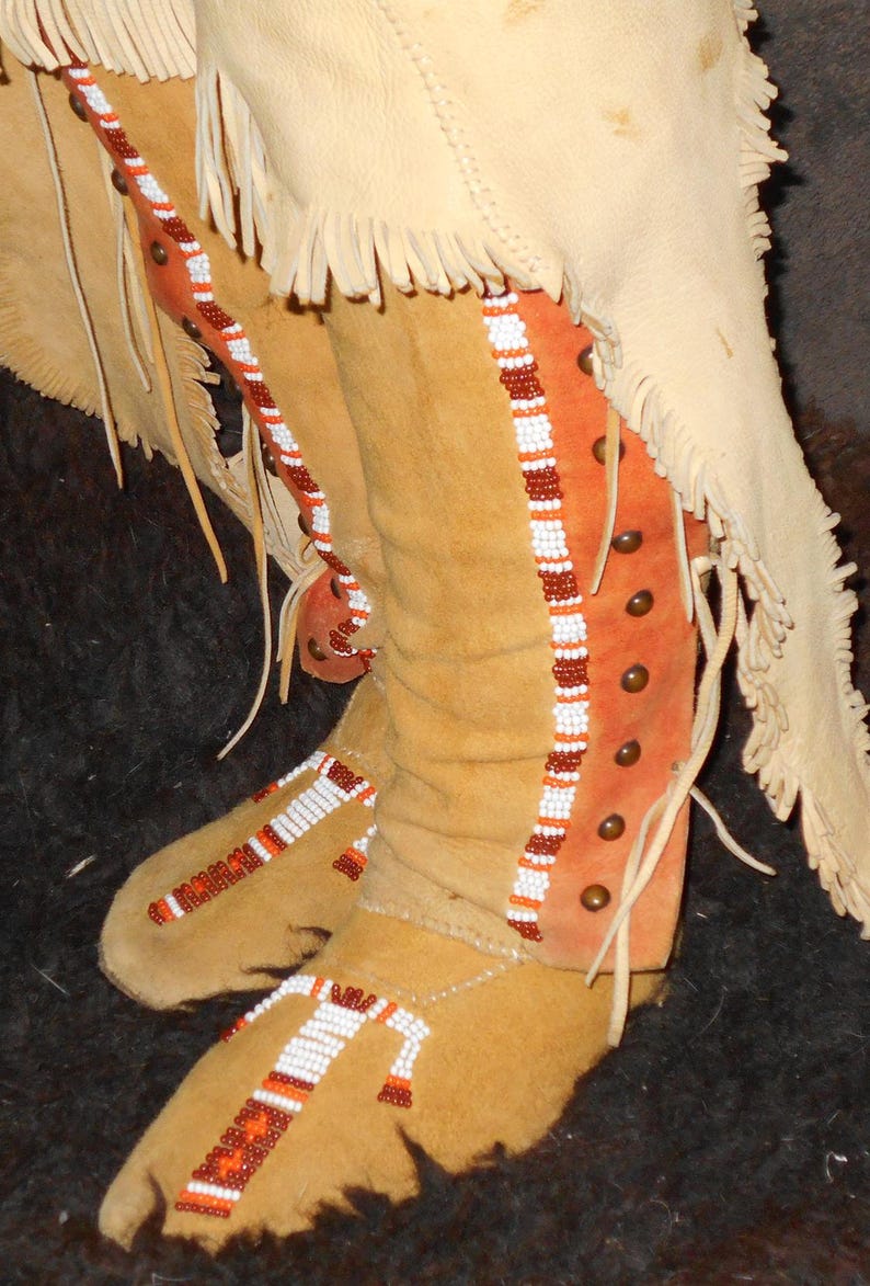 May include: Close-up of a pair of tan leather moccasins with intricate beadwork. The moccasins have a fringe detail and are decorated with orange, white, and brown beads.