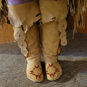 May include: A pair of tan leather moccasins with red and yellow beadwork designs. The moccasins have a fringe detail and are decorated with small round beads.