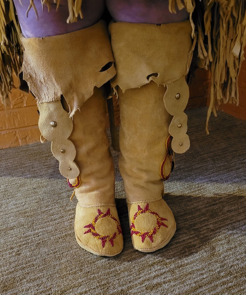 May include: A pair of tan leather moccasins with red and yellow beadwork designs. The moccasins have a fringe detail and are decorated with small round beads.