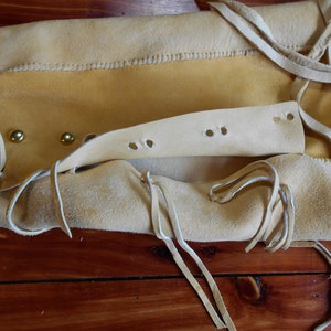 May include: Close-up of a light brown leather bag with a flap and a strap. The bag has a decorative fringe and brass rivets. The bag is open and the inside is visible.