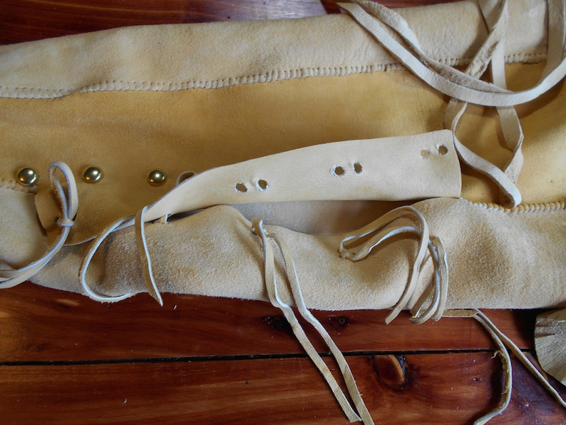 May include: Close-up of a light brown leather bag with a flap and a strap. The bag has a decorative fringe and brass rivets. The bag is open and the inside is visible.