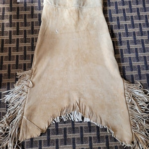 May include: A tan leather skirt with fringe detailing. The skirt has a high waist and a straight silhouette. The fringe is made of leather and is about 10 inches long.