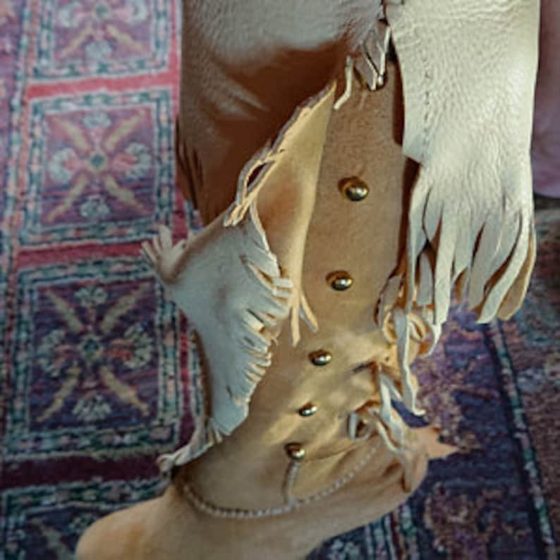 May include: Tan leather knee-high boots with fringe detail and gold studs. The boots have a lace-up front and a pointed toe.