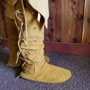 May include: A pair of tan leather moccasins with laces and metal accents. The moccasins are worn and have a worn look.