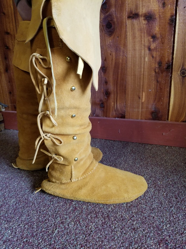 May include: A pair of tan leather moccasins with laces and metal accents. The moccasins are worn and have a worn look.