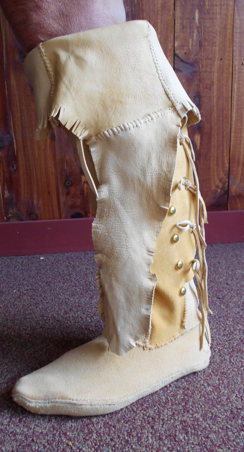 May include: A pair of tan leather moccasins with a fringe detail and a lace-up design. The moccasins have a pointed toe and a flat sole.