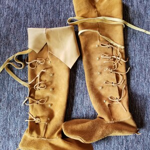 May include: A pair of brown leather knee-high boots with laces and decorative metal accents. The boots are made of a soft, supple leather and have a pointed toe.