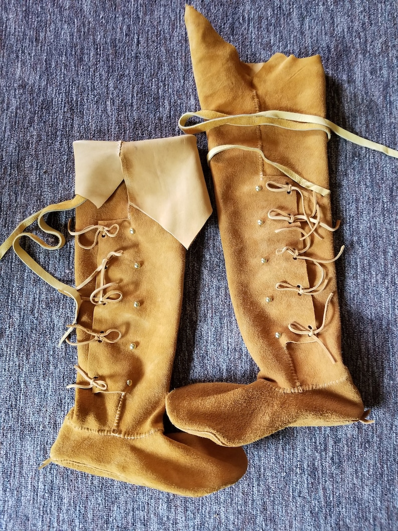 May include: A pair of brown leather knee-high boots with laces and decorative metal accents. The boots are made of a soft, supple leather and have a pointed toe.