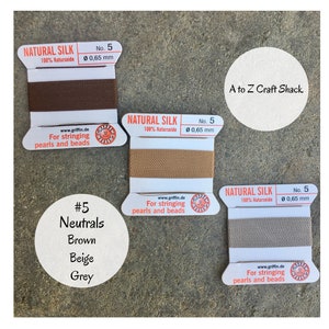 May include: Three spools of natural silk thread for stringing pearls and beads. The spools are labeled "Natural Silk 100% Naturseide" and are in the colors brown, beige, and gray. The spools are labeled with the number 5 and the diameter 0.65 mm. The text "A to Z Craft Shack" is visible in the upper right corner of the image.