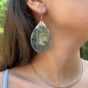 May include: Silver-toned filigree teardrop earrings with intricate floral cutouts. The earrings hang from a silver hook and are worn with a matching silver chain necklace. The earrings are a statement piece.