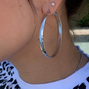 Peut inclure: Boucles d'oreilles créoles en argent avec une finition lisse et polie. Les boucles d'oreilles sont grandes et circulaires, avec un diamètre d'environ 7,5 cm.