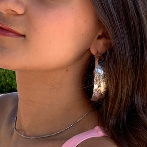 May include: A silver hoop earring with a textured surface. The earring is worn by a person with long brown hair.