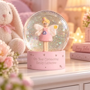 May include: A glass snow globe containing a fairy figurine with blonde hair, a pink dress, and white wings. The base is light pink with small heart icons and the engraved text "On Your Christening Emily Catherine." The globe is filled with iridescent glitter.