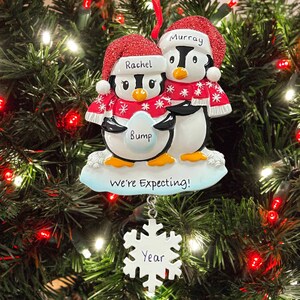 May include: A Christmas bauble featuring two penguins wearing Santa hats and scarves. The female penguin is holding a blue egg that says "Bump" and the male penguin is holding a sign that says "We're Expecting!" A snowflake bauble with the word "Year" is hanging below the penguins.