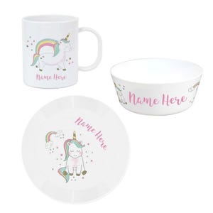 May include: A white ceramic mug, bowl, and plate set with a unicorn design, rainbow mane, and tail. Each item has the text "Name Here" in pink script. The set is designed for children.