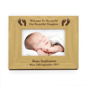 May include: A light brown wooden photo frame with a baby photo inside. The frame has an engraved message that reads "Welcome To The World Our Beautiful Daughter" with a footprint on each side. The name "Rosie Stephenson" and the date "Born 29th September 2017" are engraved on the bottom of the frame.