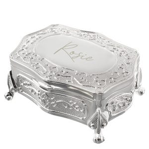 May include: A silver-colored jewelry box with ornate floral detailing and four small feet. The lid is engraved with the name "Rosie" in cursive script. The box has a rectangular shape with rounded edges.