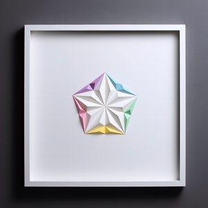 May include: A framed, geometric paper art piece featuring a white, multi-faceted star with pastel-colored tips. The star is centered on a white background within a white square frame, set against a gray backdrop.