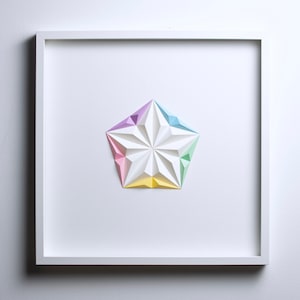 May include: A framed, geometric paper art piece featuring a white, multi-faceted star shape with pastel color accents. The star is composed of folded paper, with sections of pink, yellow, blue, and purple. The artwork is set against a white background.