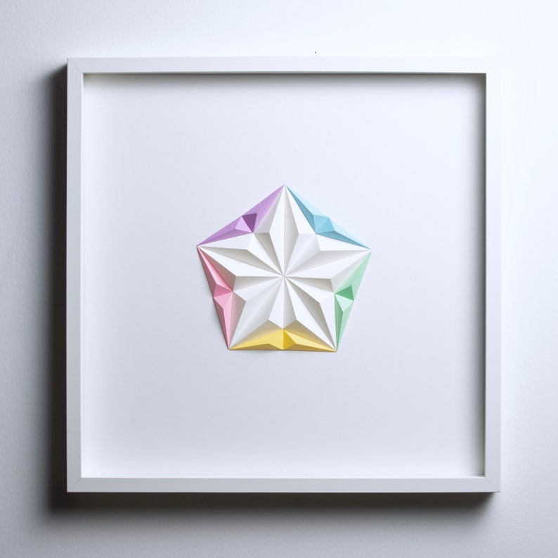 May include: A framed, geometric paper art piece featuring a white, multi-faceted star shape with pastel color accents. The star is composed of folded paper, with sections of pink, yellow, blue, and purple. The artwork is set against a white background.