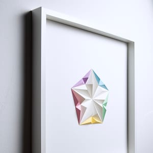 May include: A framed artwork featuring a geometric paper sculpture in pastel colors. The white frame surrounds a white background with a multi-faceted star-like shape in shades of pink, purple, blue, green, and yellow. The artwork is mounted on a white wall.