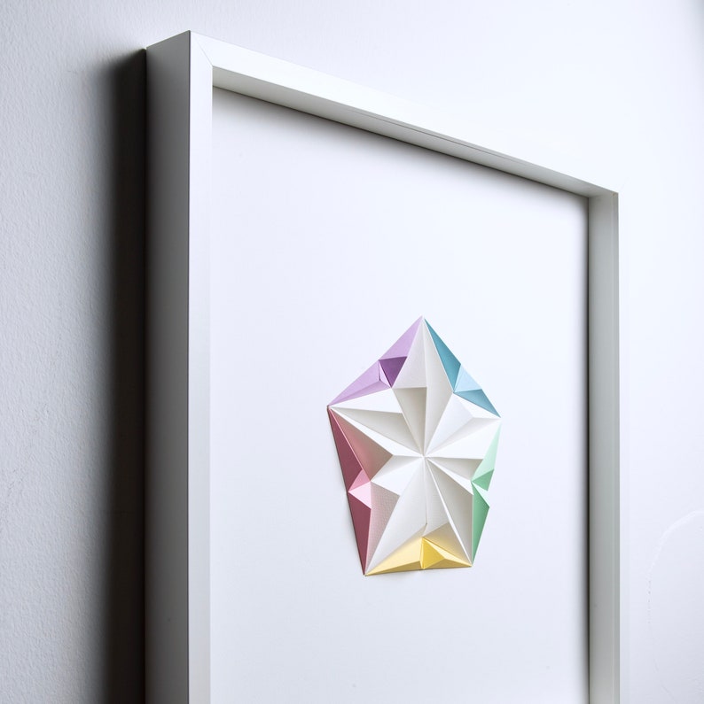 May include: A framed artwork featuring a geometric paper sculpture in pastel colors. The white frame surrounds a white background with a multi-faceted star-like shape in shades of pink, purple, blue, green, and yellow. The artwork is mounted on a white wall.