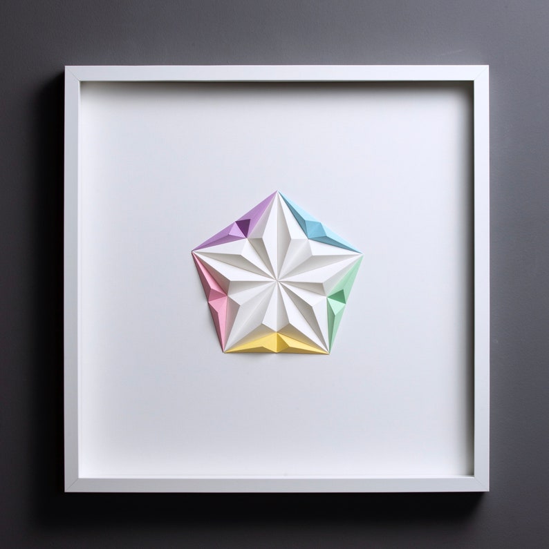 May include: A framed, geometric paper art piece featuring a white, multi-faceted star with pastel-colored tips. The star is centered on a white background within a white square frame, set against a gray backdrop.