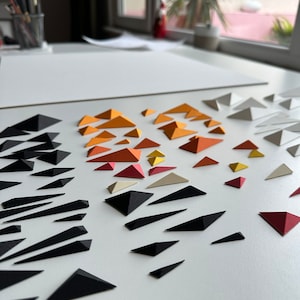 Puede incluir: Una superficie blanca con una variedad de tri&aacute;ngulos de papel en diferentes colores, incluyendo negro, blanco, rojo, naranja y amarillo. Los tri&aacute;ngulos est&aacute;n dispuestos en un patr&oacute;n, con los tri&aacute;ngulos negros formando una l&iacute;nea diagonal a trav&eacute;s de la superficie.