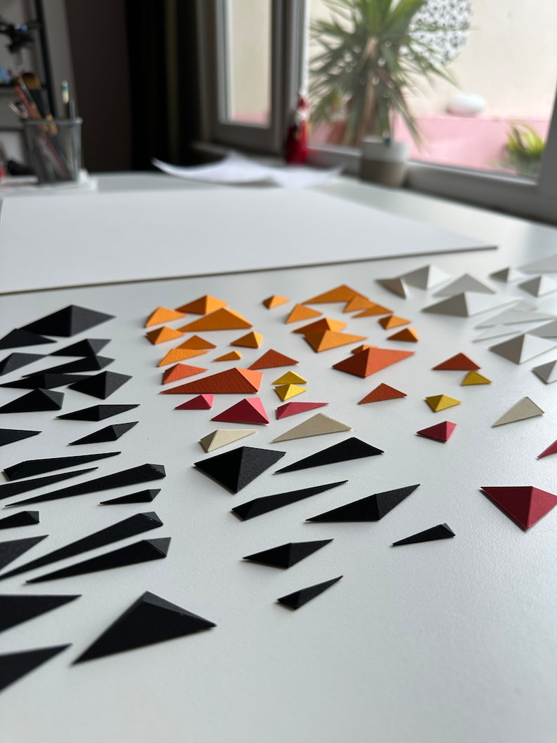 Puede incluir: Una superficie blanca con una variedad de tri&aacute;ngulos de papel en diferentes colores, incluyendo negro, blanco, rojo, naranja y amarillo. Los tri&aacute;ngulos est&aacute;n dispuestos en un patr&oacute;n, con los tri&aacute;ngulos negros formando una l&iacute;nea diagonal a trav&eacute;s de la superficie.