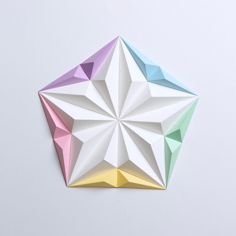 May include: A geometric paper art piece in the shape of a five-pointed star. The star is composed of white, pastel pink, yellow, green, light blue, and purple paper. The star's design features a series of folded, triangular facets.