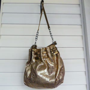 May include: A metallic gold bucket bag with a textured snakeskin pattern. The bag features a long shoulder strap, silver-toned metal chain links, and decorative horizontal straps with silver buckles around the upper portion of the bag.