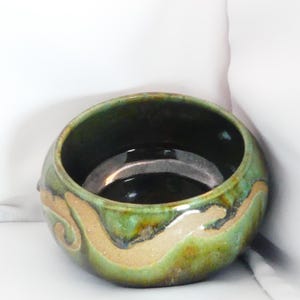 May include: A round ceramic bowl with a glossy finish, featuring a deep green and brown glaze. The exterior displays a swirling, unglazed tan clay band that wraps around the middle, contrasting with the dark, reflective interior.