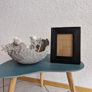 Puede incluir: Un cuenco de hormig&oacute;n gris con flores secas blancas se encuentra sobre una mesa azul verdosa con patas de madera. Un marco de imagen negro con un paspart&uacute; marr&oacute;n est&aacute; sobre la mesa.
