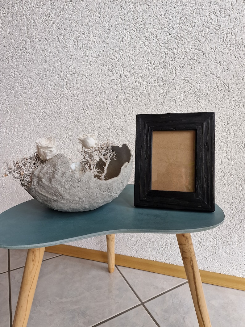 Puede incluir: Un cuenco de hormig&oacute;n gris con flores secas blancas se encuentra sobre una mesa azul verdosa con patas de madera. Un marco de imagen negro con un paspart&uacute; marr&oacute;n est&aacute; sobre la mesa.