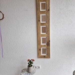 Pu&ograve; includere: Una cornice per foto in legno rustico con cinque aperture per foto. La cornice &egrave; realizzata in legno riciclato e ha una finitura naturale. La cornice &egrave; appesa a una parete bianca.