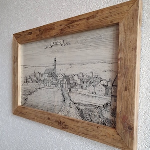 Puede incluir: Un dibujo en blanco y negro enmarcado de una ciudad hist&oacute;rica con la palabra "Roding" en una pancarta en la parte superior. El dibujo representa una ciudad con un r&iacute;o que la atraviesa y una iglesia con un alto campanario en el centro.
