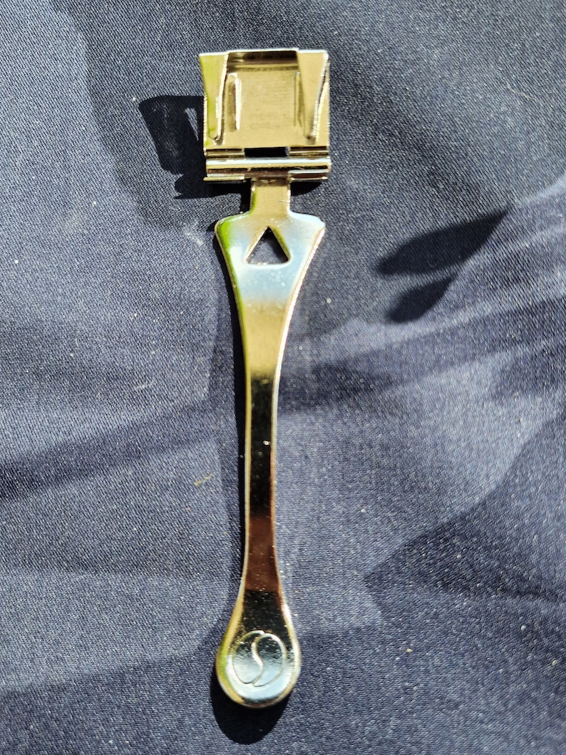 Puede incluir: Un cierre de pulsera de reloj de metal plateado con un dise&ntilde;o triangular y un extremo redondeado. El cierre tiene un peque&ntilde;o logotipo grabado en el extremo.