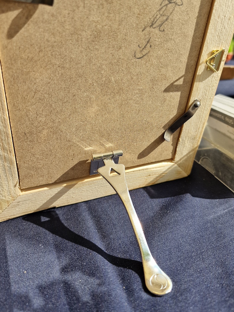 Puede incluir: Un marco de fotos de madera con un caballete de metal plateado. El marco tiene un acabado marr&oacute;n claro y un dise&ntilde;o sencillo.