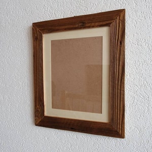 May include: A rustic wooden picture frame with a light brown wood finish. The frame has a white mat and is hanging on a white textured wall.