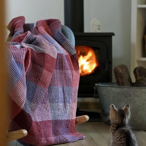 Puede incluir: Una escena acogedora con una manta a cuadros roja, morada y azul sobre una mecedora. Una chimenea encendida y un pequeño gatito atigrado en el suelo de madera contribuyen al ambiente.