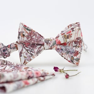 Könnte beinhalten: Ein weißer Fliege mit einem floralen Muster in Rosa-, Braun- und Grüntönen. Die Fliege wird mit einem passenden Einstecktuch geliefert.