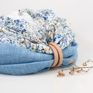 May include: A round, blue fabric container with a floral patterned top. The container is secured with two light brown leather straps. The floral pattern features blue, yellow, and white flowers. A small sprig of dried flowers is in the foreground.
