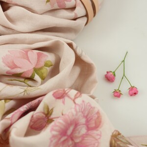 May include: A pale pink linen scarf with a floral print of pink roses and green leaves. The scarf is folded and tied with a brown leather strap. A small bouquet of pink flowers is in the bottom right corner of the image.