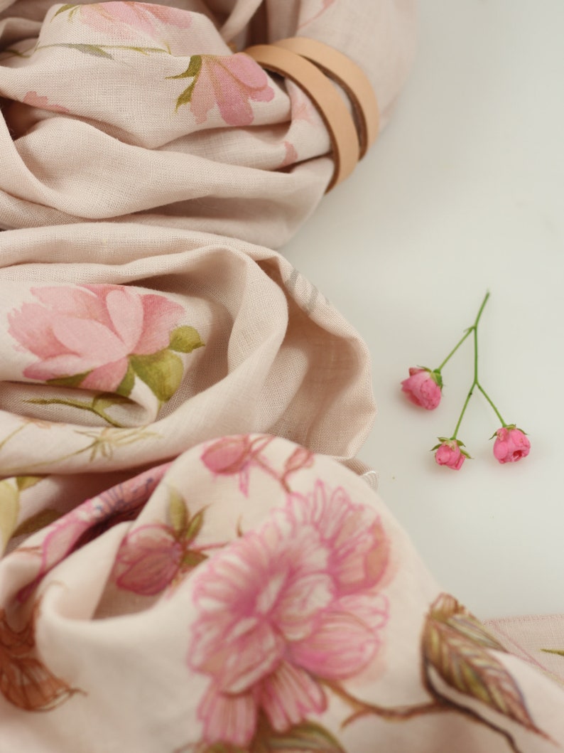 May include: A pale pink linen scarf with a floral print of pink roses and green leaves. The scarf is folded and tied with a brown leather strap. A small bouquet of pink flowers is in the bottom right corner of the image.