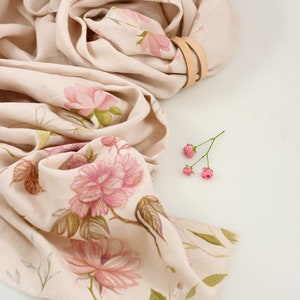 May include: A beige linen scarf with a floral print of pink flowers and green leaves. The scarf is folded and tied with a brown leather strap.