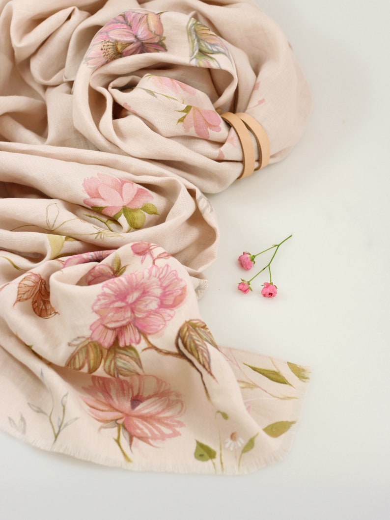 May include: A beige linen scarf with a floral print of pink flowers and green leaves. The scarf is folded and tied with a brown leather strap.