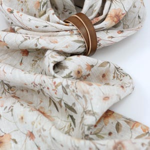 May include: A white linen scarf with a floral print in shades of brown, orange, and green. The scarf is tied in a knot and has a brown leather strap around it.