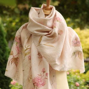May include: A pale pink silk scarf with a floral print of pink peonies and green leaves. The scarf is draped over a mannequin.