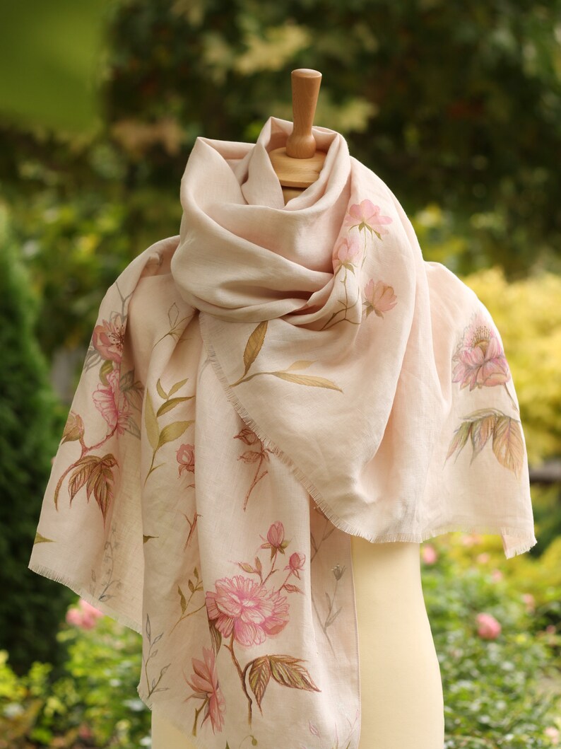 May include: A pale pink silk scarf with a floral print of pink peonies and green leaves. The scarf is draped over a mannequin.