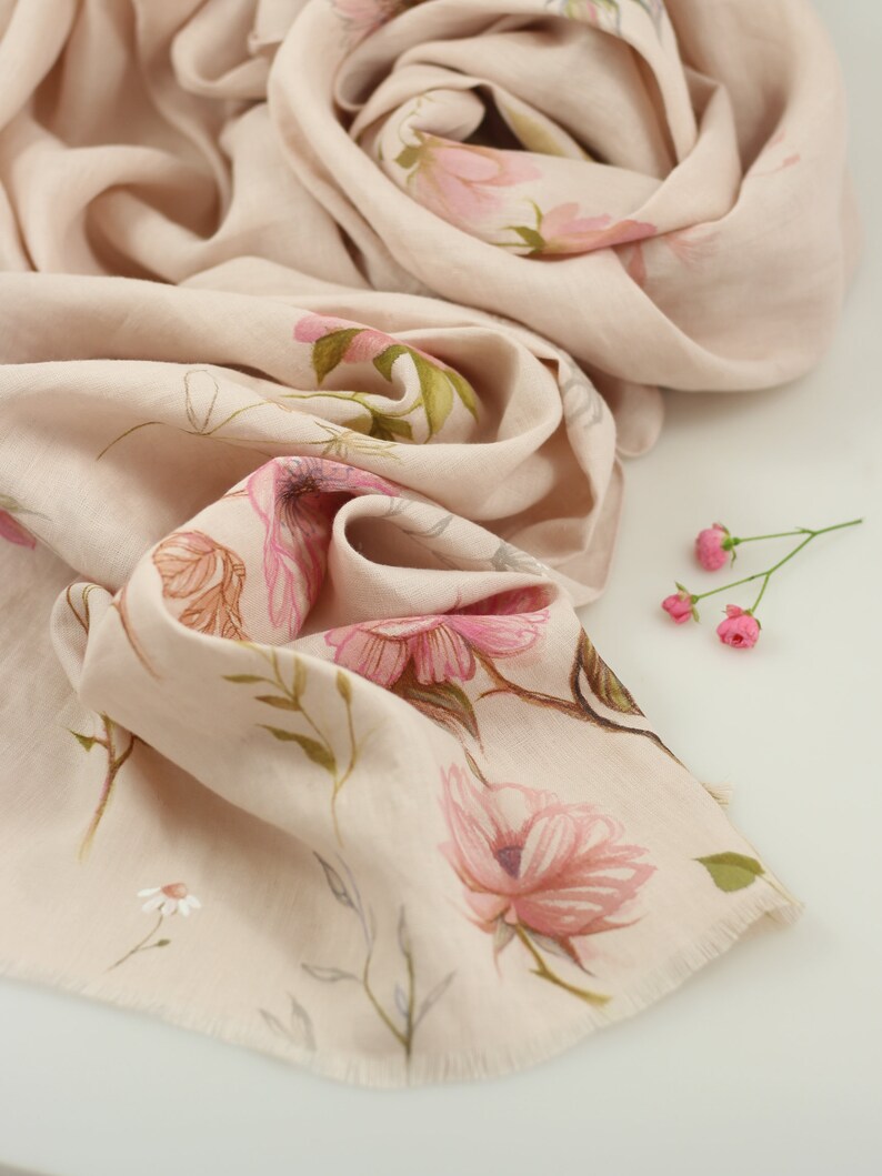 May include: A soft, blush-colored scarf with a delicate floral print. The scarf is made of a lightweight fabric and has a fringed edge.